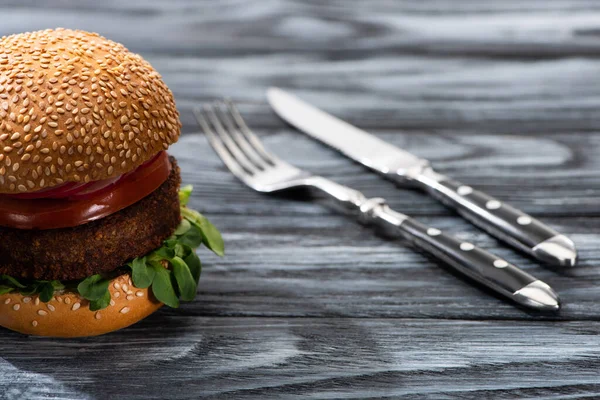 Foco seletivo de hambúrguer vegan saboroso com legumes servidos em mesa de madeira com garfo e faca — Fotografia de Stock