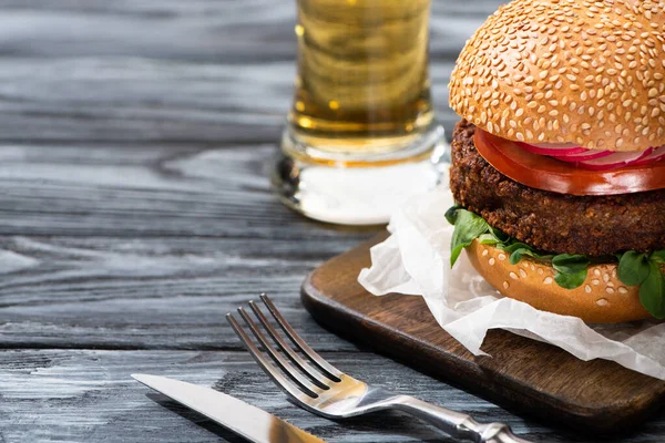 Selektiver Schwerpunkt leckerer veganer Burger auf Holztisch mit Bier und Besteck — Stockfoto