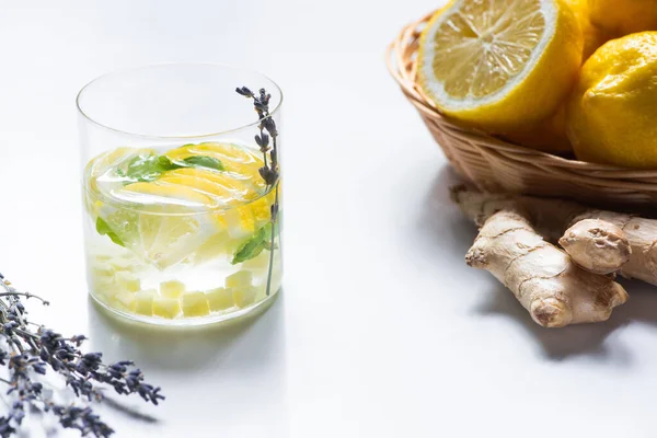 Limonata fresca allo zenzero in vetro con rametto di menta e lavanda vicino cesto di limoni e radice di zenzero su fondo bianco — Foto stock