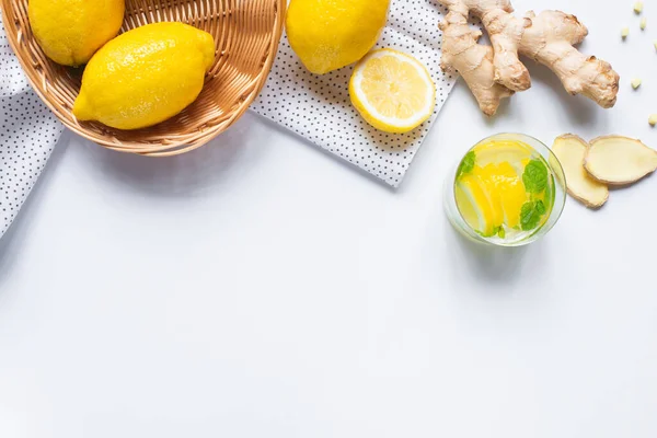Вид на свежий лимонад в стакане возле корзины с лимонами и корень имбиря на белом фоне с салфеткой — Stock Photo