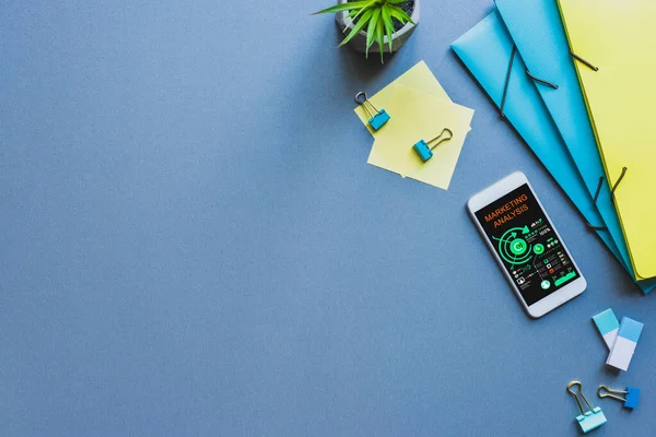 Vista superior del teléfono inteligente con aplicación de análisis de marketing cerca de papelería en la superficie azul - foto de stock