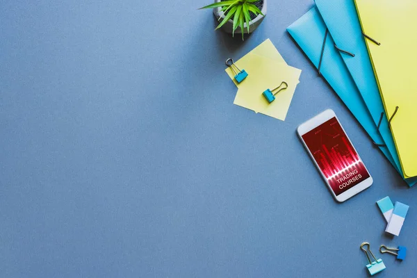 Top view of stationery and smartphone with trading courses app on blue surface — Stock Photo