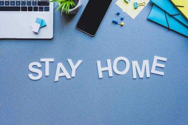 Top view of lettering stay home near smartphone, laptop and stationery on blue surface — Stock Photo