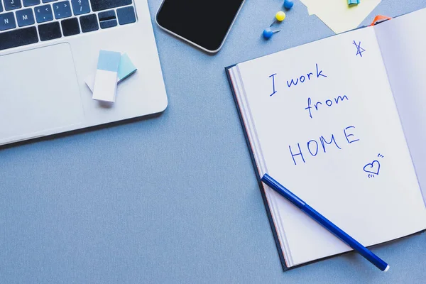 Vista superior de gadgets, suministros de oficina y letras que trabajo desde casa en el cuaderno abierto en la superficie azul - foto de stock