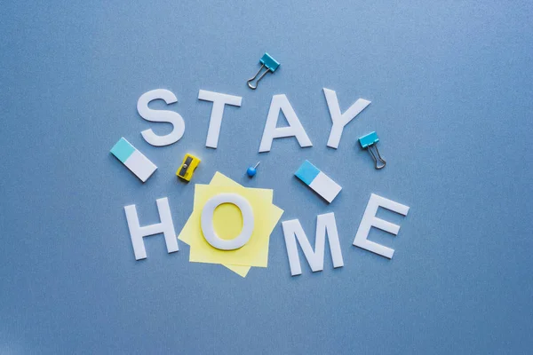 Top view of stay home lettering near sticky notes, binder clips and pencil sharpener on blue surface — Stock Photo