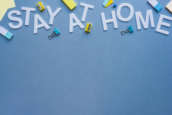 Top view of stay at home lettering near pencil sharpeners, erasers and binder clips on blue surface — Stock Photo