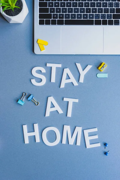 Top view of stay at home lettering near laptop, plant and stationery on blue background — Stock Photo