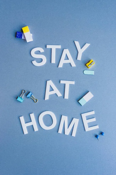 Top view of stay at home lettering near erasers, binder clips and pencil sharpener on blue surface — Stock Photo