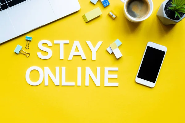 Vista superior de ficar online lettering perto de dispositivos digitais, xícara de café e papelaria na superfície amarela — Fotografia de Stock