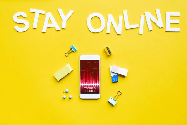 Top view of smartphone with trading courses app, office supplies and stay online lettering on yellow surface — Stock Photo