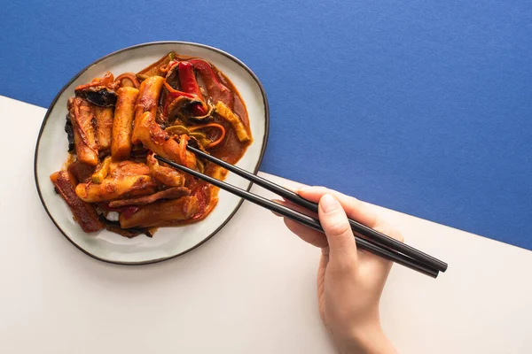 Cropped view of woman holding chopsticks near korean topokki with sauce on white and blue — Stock Photo