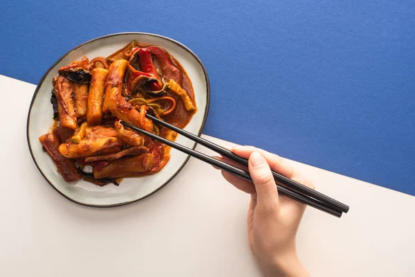 Vue recadrée de femme tenant des baguettes près de topokki coréen avec sauce sur blanc et bleu — Photo de stock