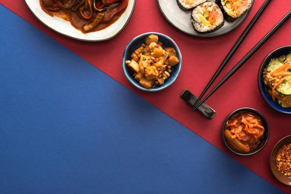 Top view of bowls and plates with spicy korean kimchi and kimbap on blue and crimson — Stock Photo