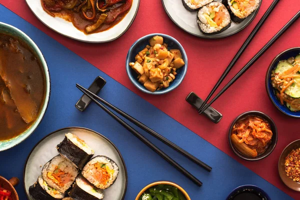 Vista superior de pauzinhos perto de ramen, kimbap e pratos laterais coreanos em azul e carmesim — Fotografia de Stock