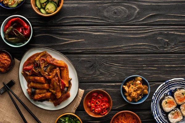 Vue de dessus de topokki coréen près de kimbap, kimchi, baguettes, plats d'accompagnement et serviette en coton sur la surface en bois — Photo de stock