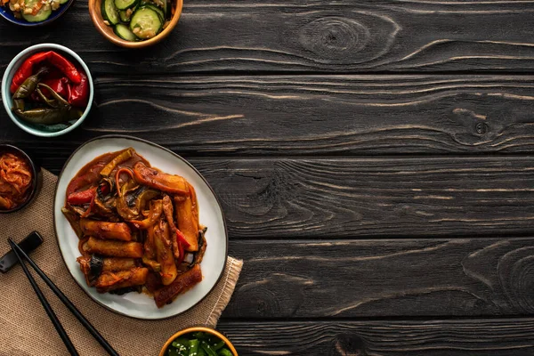 Vue de dessus de topokki coréen près de kimchi, baguettes, plats d'accompagnement et serviette en coton sur la surface en bois — Photo de stock