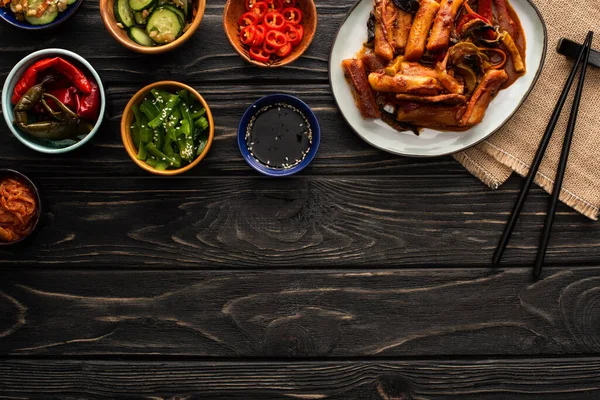 Vue de dessus de topokki coréen près de baguettes, plats d'accompagnement savoureux et serviette en coton sur la surface en bois — Photo de stock