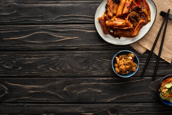 Vista dall'alto di topokki coreani piccanti vicino a patate brasate, cetrioli sottaceto, bacchette e tovagliolo di cotone sulla superficie di legno — Foto stock