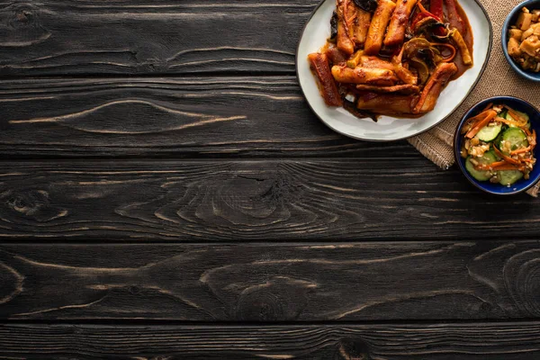 Draufsicht auf würzige Topokki in der Nähe koreanischer Schmorkartoffeln, eingelegter Gurken, Essstäbchen und Baumwollserviette auf Holzoberfläche — Stockfoto