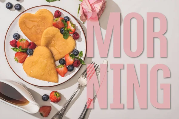 Top view of delicious heart shaped pancakes with berries near rose, cutlery and maple syrup on white background, morning illustration — Stock Photo