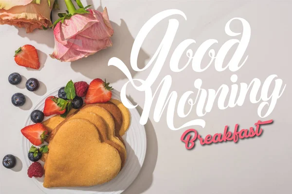 Vista superior de deliciosas panquecas em forma de coração com bagas perto de rosas no fundo branco, boa ilustração matinal — Fotografia de Stock