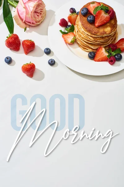 Top view of delicious pancakes with blueberries and strawberries on plate near rose flower on marble white surface, good morning illustration — Stock Photo