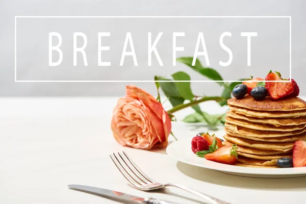 Selective focus of delicious pancakes with blueberries and strawberries on plate near rose flower and cutlery on white surface isolated on grey, breakfast illustration — Stock Photo