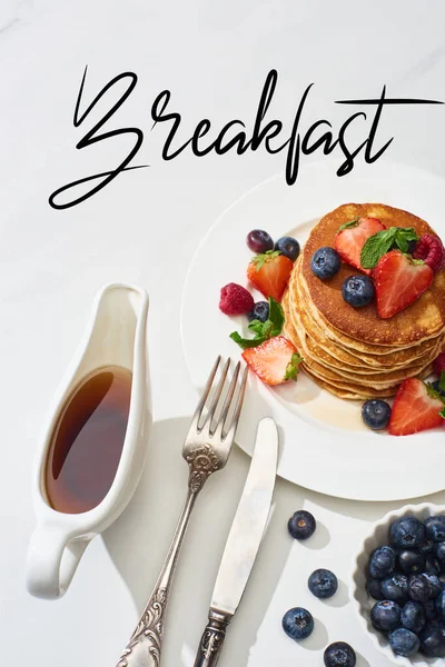 Vista superior de deliciosos panqueques con jarabe de arce, arándanos y fresas en el plato cerca de tenedor y cuchillo en la superficie blanca de mármol, ilustración del desayuno — Stock Photo