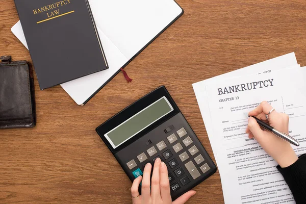 Partial view of woman filling in bankruptcy form and using calculator on wooden background — Stock Photo