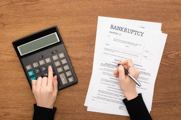 Cropped view of woman filling in bankruptcy form and using calculator on wooden background — Stock Photo