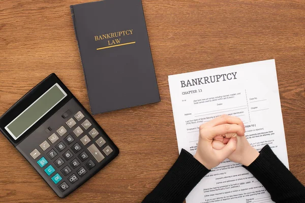 Vista recortada de las manos apretadas hembra en papel de bancarrota cerca de libro de leyes de bancarrota y calculadora sobre fondo de madera - foto de stock