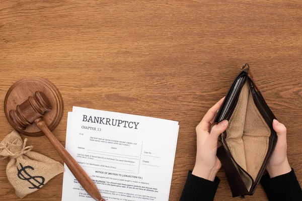 Cropped view of woman holding empty wallet near bankruptcy documents, money bag and gavel on wooden background — Stock Photo