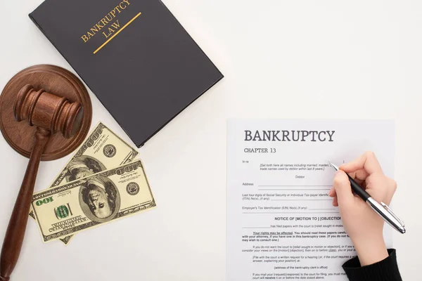 Vista recortada de la mujer llenando el formulario de bancarrota con pluma cerca de dinero, mazo y libro de leyes de bancarrota sobre fondo blanco - foto de stock