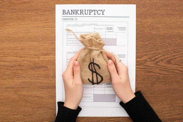 Cropped view of woman holding money bag near bankruptcy paper on wooden background — Stock Photo