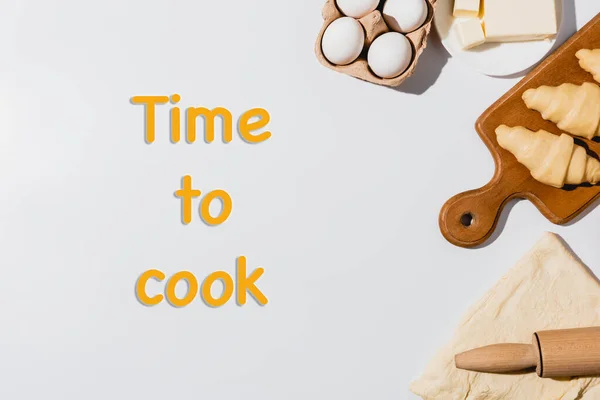 Top view of fresh raw croissants on wooden cutting board near rolling pin, butter and eggs on white background with time to cook lettering — Stock Photo