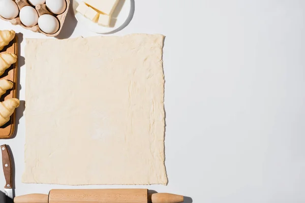 Вид сверху на свежие круассаны на деревянной разделочной доске рядом с сырым тестом, нож, скалка, масло и яйца на белом фоне — стоковое фото