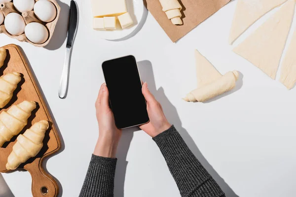 Vista recortada de la mujer sosteniendo teléfono inteligente con pantalla en blanco mientras cocina croissant sobre fondo blanco - foto de stock