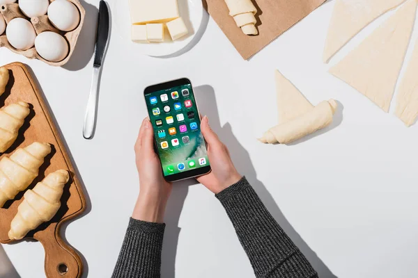 KYIV, UCRANIA - 22 DE ABRIL DE 2019: vista recortada de la mujer sosteniendo el iPhone mientras cocina croissant sobre fondo blanco - foto de stock