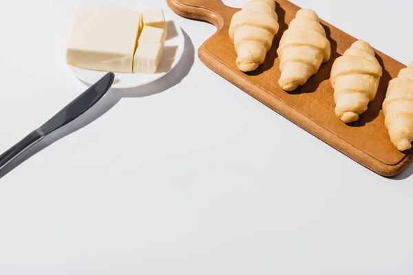 Cruasanes crudos frescos sobre tabla de cortar de madera cerca de mantequilla en plato con cuchillo sobre fondo blanco - foto de stock