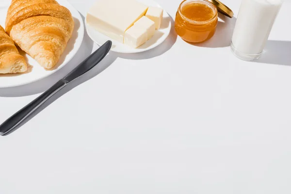 Вкусные свежие круассаны на тарелке возле стакана молока, масло с ножом и варенье на белом фоне — Stock Photo