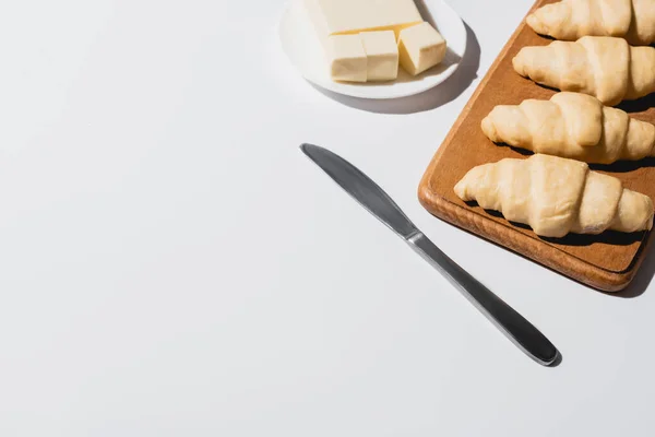 Fresh raw croissants on wooden cutting board near butter on plate with knife on white background — Stock Photo