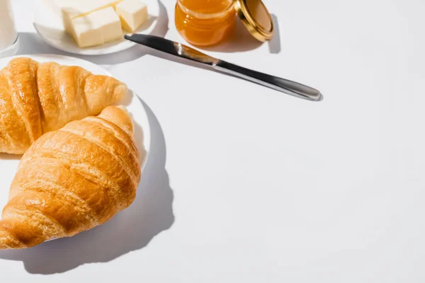 Saborosos croissants frescos assados na placa perto de manteiga com faca e geléia no fundo branco — Fotografia de Stock