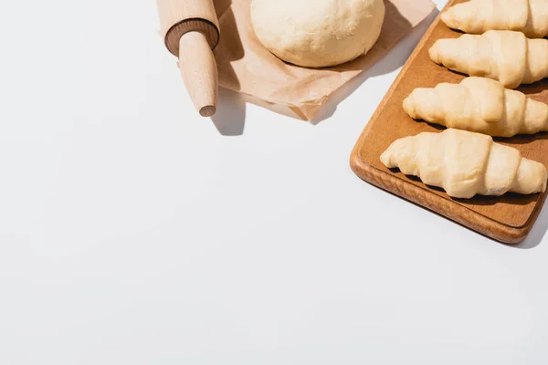 Fresh croissants on wooden cutting board near raw dough, rolling pin on white background — Stock Photo