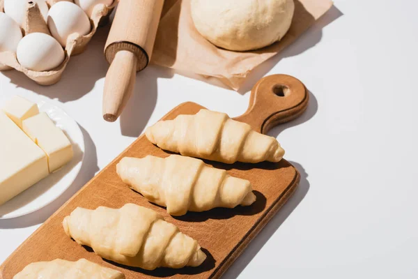 Frische Croissants auf Holzschneidebrett in der Nähe von rohem Teig, Nudelholz, Butter und Eiern auf weißem Hintergrund — Stockfoto