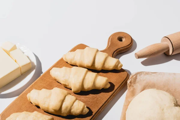 Croissants frais sur planche à découper en bois près de pâte crue, rouleau à pâtisserie, beurre sur fond blanc — Photo de stock