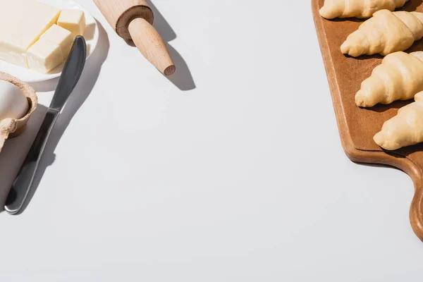 Свежие круассаны на деревянной разделочной доске возле ножа, скалка, масло на белом фоне — стоковое фото