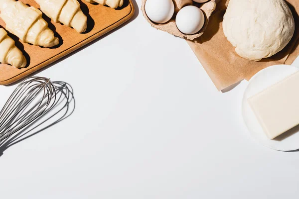 Top view of fresh croissants on wooden cutting board near raw dough, balloon whisk, butter and eggs on white background — Stock Photo