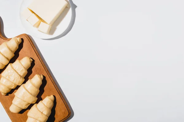 Top view of fresh raw croissants on wooden cutting board near butter on plate on white background — Stock Photo