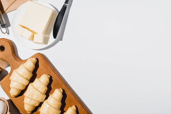 Top view of fresh raw croissants on wooden cutting board near butter on plate with knife on white background — Stock Photo