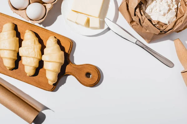 Vista superior de croissants crus na placa de corte de madeira perto de ingredientes no fundo branco — Fotografia de Stock